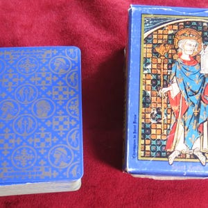 K&ouml;nnte beinhalten: Ein blaues Kartenspiel mit einem gold-blauen Muster. Die Schachtel zeigt ein Buntglasbild einer Person in einem blauen und roten Gewand, die einen Stab und ein kleines Geb&auml;ude h&auml;lt.