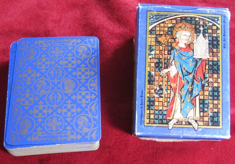 K&ouml;nnte beinhalten: Ein blaues Kartenspiel mit einem gold-blauen Muster. Die Schachtel zeigt ein Buntglasbild einer Person in einem blauen und roten Gewand, die einen Stab und ein kleines Geb&auml;ude h&auml;lt.