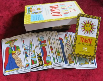 Könnte beinhalten: Ein Tarot-Kartendeck mit den großen Arkana, das auf einer roten Oberfläche ausgelegt ist. Die Karten zeigen farbenfrohe Illustrationen und französischen Text. Die Schachtel für die Karten ist braun mit goldenen Schriftzügen und einer Sonnenillustration. Der Text auf der Schachtel lautet "Tarot de Marseille 22 arcanes majeurs".