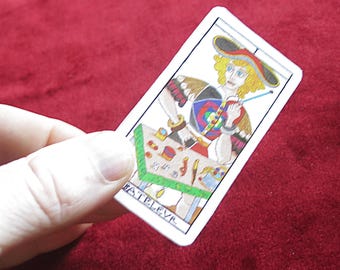 May include: A tarot card featuring a colorful illustration of a person with a hat, holding a tool, and surrounded by various objects. The card has a white border and the word "FATELEVR" is visible.