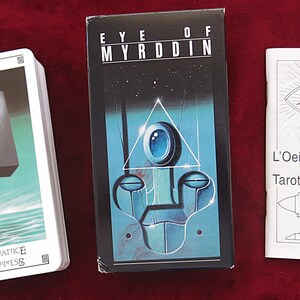 May include: A tarot card deck and accompanying booklet are displayed on a red surface. The deck box is black with the title "EYE OF MYRDDIN". A deck of cards shows "L'IMPERATRICE THE EMPRESS". The booklet is titled "L'Oeil de Myrddin ou Tarot des Princes".