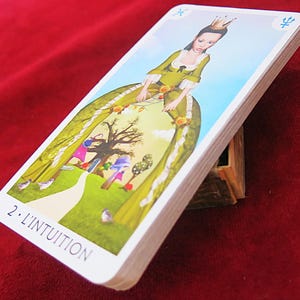 May include: A deck of tarot cards, titled "2. L'INTUITION", is propped up on a wooden stand. The card on top features a woman in a green dress with a crown, set against a landscape with a path and trees. The background is a deep red.