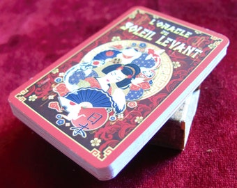 Puede incluir: Una carta roja y dorada con el texto "L'Oracle du Soleil Levant" y una ilustración colorida de una mujer con un kimono japonés tradicional sosteniendo un abanico.