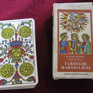 May include: A deck of Tarot cards and its box. The cards feature colorful illustrations with yellow, blue, and red accents. The box has a brown label with the text "TAROCCHI MARSIGLIESI" and illustrations of people and an angel.