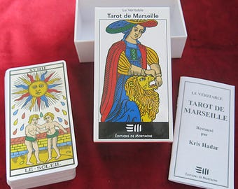May include: A deck of tarot cards with a white background and colorful illustrations. The deck is titled "Le Véritable Tarot de Marseille" and is restored by Kris Hadar. The deck is in a white box with the same title and a smaller booklet with the same title.