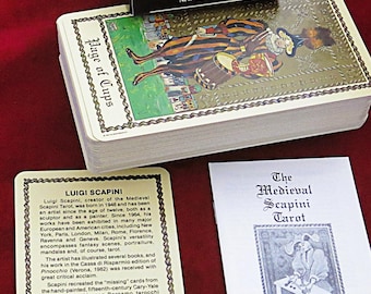May include: A tarot card deck with a black box labeled "The Medieval Scapini Tarot." The deck includes cards with medieval-themed illustrations and text. A booklet and a card with text are also visible.