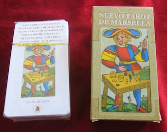 May include: A deck of tarot cards in a white box with the title "Nuevo Tarot de Marsella" in gold lettering. The box is wrapped in plastic and has a yellow ribbon. The cards feature a colourful illustration of a person in a red, yellow, and blue costume holding a knife and a stick. The illustration is surrounded by a white border.