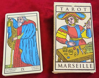 May include: A Tarot de Marseille deck featuring a box with a colorful illustration of a figure at a table and a single card showing The Hermit, labeled L'ERMITE, IX, EL ERMITAÑO THE HERMIT, and L'ERMITA O ERMITA in black text.
