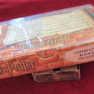 Puede incluir: Una caja de cartón marrón con una cubierta de envoltura de plástico. La caja tiene un patrón de ladrillo y el texto "Harry Potter" impreso en la parte delantera. El texto "Folleto de instrucciones incluido" está impreso en la esquina inferior derecha de la caja.