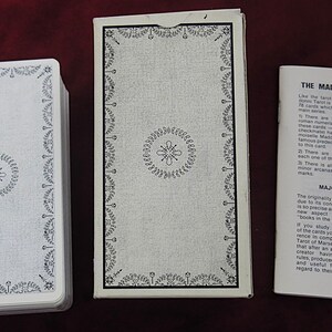 May include: A tarot card deck with a white and black design. The deck, box, and booklet are displayed on a dark red surface. The box and card backs feature a floral border and central emblem. The booklet is titled "THE MADDONNI TAROT".