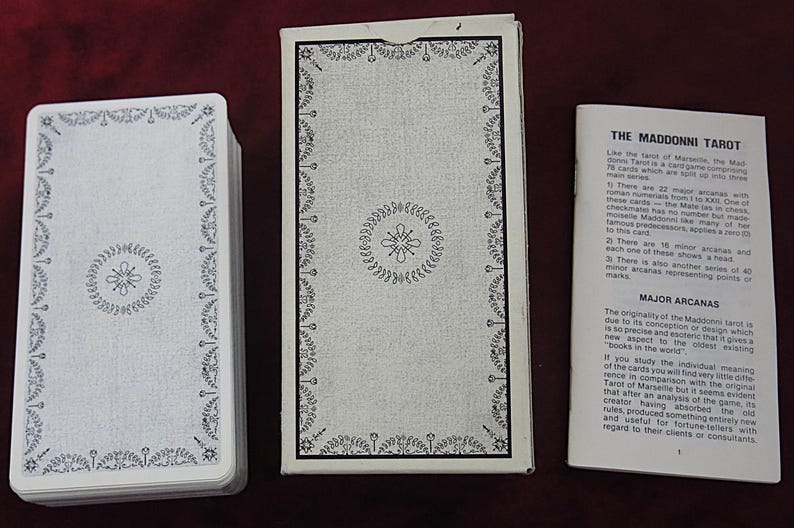 May include: A tarot card deck with a white and black design. The deck, box, and booklet are displayed on a dark red surface. The box and card backs feature a floral border and central emblem. The booklet is titled "THE MADDONNI TAROT".