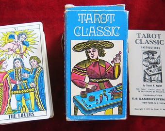 May include: A vintage Tarot Classic deck with a blue box featuring a detailed illustration. The deck includes a card stack with "THE LOVERS" and a booklet. The box and booklet have the text "TAROT CLASSIC". The cards and box are on a red surface.