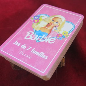 May include: A pink card game box featuring a picture of Barbie on the front. The box reads "Barbie Jeu de 7 familles Ducale".