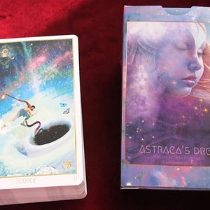 May include: A deck of tarot cards and its box titled Astraca's Dream. The card on top shows a figure surfing on a wave toward a cup of dark liquid under a starry sky. The box features a large, ethereal face with closed eyes and cosmic colors.