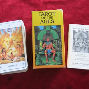 Op de afbeelding: Een tarotdeck doos met een gele achtergrond en de tekst "Tarot of the Ages". De doos heeft een illustratie van een persoon met een hoofdtooi die op een troon zit. Het kaartspel is zichtbaar op de achtergrond met de kaart "Jugement" met de afbeelding naar boven.