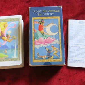 Op de afbeelding: Een tarotdeck doos met een blauwe achtergrond en een vrouw die op een lotusbloem zit. De doos is gelabeld "Tarot du Voyage en Orient". Het deck bevat 78 kaarten met illustraties van de grote en kleine arcana.