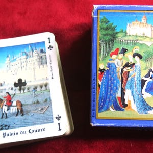 K&ouml;nnte beinhalten: Ein Kartenspiel mit einem Bild des Louvre-Palastes auf der Karte As der Kreuz. Die Schachtel zeigt eine mittelalterliche Szene mit Personen in bunten Kleidern.