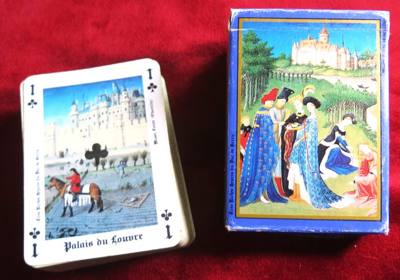 K&ouml;nnte beinhalten: Ein Kartenspiel mit einem Bild des Louvre-Palastes auf der Karte As der Kreuz. Die Schachtel zeigt eine mittelalterliche Szene mit Personen in bunten Kleidern.