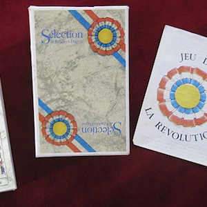 Könnte beinhalten: Eine Sammlung von Vintage-Spielkarten und -schachteln. Die Schachteln zeigen den Text "Selection" und ein Rosettendesign in Rot, Weiß und Blau. Eine Karte zeigt einen Engel mit einem Schild. Die Karten liegen auf rotem Untergrund.