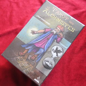 May include: A boxed tarot deck titled "Le Tarot des Alchimistes" by Jean Beauchard. The box features an illustration of an alchemist in a robe, with a thought bubble showing a bird and a wolf. The background is a mix of brown and blue.