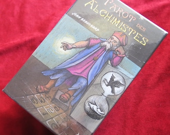 May include: A boxed tarot deck titled "Le Tarot des Alchimistes" by Jean Beauchard. The box features an illustration of an alchemist in a robe, with a thought bubble showing a bird and a wolf. The background is a mix of brown and blue.