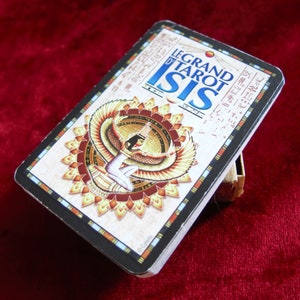 Könnte beinhalten: Ein Tarotkartenspiel mit dem Titel "Le Grand Tarot Isis" mit einer Illustration einer geflügelten Figur in einem Sonnenstrahl-Design. Die Karten haben einen schwarzen Rand und liegen auf einem kleinen, hellen Ständer vor rotem Hintergrund.