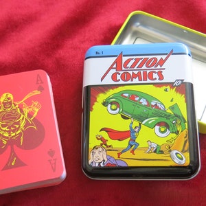 Peut inclure: Un jeu de cartes rouge avec Superman en costume jaune et rouge, avec l'As de Pique visible. Les cartes sont dans une boîte en métal avec un couvercle jaune et une image bleue et jaune de la couverture d'Action Comics #1.