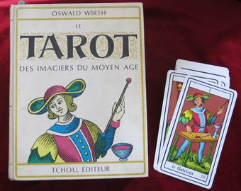 May include: A vintage book titled "Le Tarot Des Imagers Du Moyen Age" by Oswald Wirth, featuring a colorful illustration of a person holding a wand. The book is next to a tarot card with a colorful illustration of a person holding a wand and a cup. The card is labeled "Le Gateleur" with a Hebrew letter.
