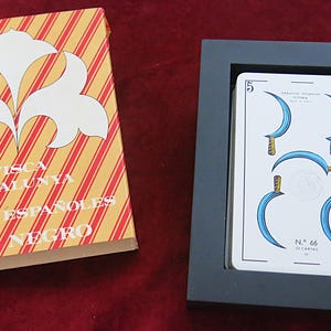 Puede incluir: Una baraja de cartas en un marco negro junto a una caja de rayas amarillas y rojas. La caja presenta gráficos florales blancos y el texto Visca Catalunya Naipes Españoles Dal Negro. La carta superior muestra un 5 y cinco hojas curvas azules.
