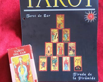 May include: A Tarot deck and guide featuring the text "Tarot de Zar" and "Tirada de la Pirámide". The deck is in a red box with a religious illustration. The guide has a black cover with yellow text and a pyramid arrangement of tarot cards.