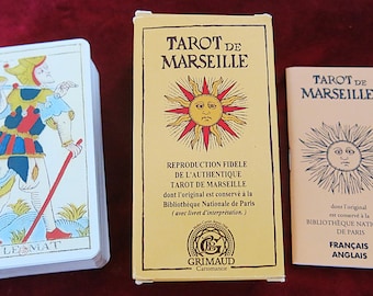 May include: A Tarot de Marseille deck featuring a Le Mat card on top, a rectangular yellow box with a sun illustration, and a small instructional booklet. The box and booklet both display the text Tarot de Marseille and a sun graphic.