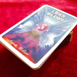 Puede incluir: Una baraja de cartas de oráculo con un diseño azul, morado y rojo que presenta un ángel con alas y una gema central. El texto "L'Oracle des Anges" y "Angela McGerr" son visibles en la carta.