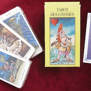 Op de afbeelding: Verschillende tarotkaartendecks worden getoond op een rode ondergrond. Eén deck zit in een gele doos met de tekst "TAROT DES GNOMES" en een illustratie van een kabouter. Andere decks zijn in plastic verpakt.