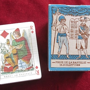 May include: A deck of playing cards and its box. The card shows a seated figure with wings, a red hat, and green clothing. The box is blue and white, with illustrations of figures and the text "PRISE DE LA BASTILLE 14 JUILLET 1789."
