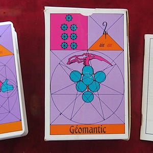Peut inclure: Un jeu de cartes de tarot Géomantique vintage avec une pile de cartes, une boîte et un livret. Les cartes et la boîte présentent des motifs géométriques en rose, violet et sarcelle, avec du texte en français. Le livret est blanc et porte le titre "Géomantique".