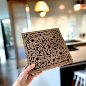 Puede incluir: Una caja de madera cuadrada con intrincados diseños florales cortados con láser. La caja se sostiene en una mano, mostrando el detallado diseño calado. La madera es de color marrón claro natural, y el interior parece oscuro.