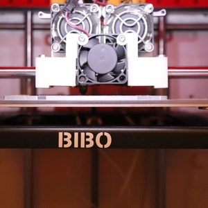 May include: Close-up of a 3D printer with a black and white fan and a black platform with the word "BIBO" printed on it.