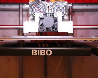 Op de afbeelding: Close-up van een 3D-printer met een zwart-witte ventilator en een zwart platform met het woord "BIBO" erop gedrukt.
