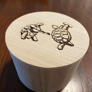 May include: A cylindrical wooden box with a lid featuring two turtles etched into the wood. One turtle appears to be playing a banjo. The box is made of light-colored wood and sits on a dark wooden surface.