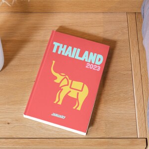 以下が含まれることがあります： 「THAILAND 2023」というタイトルと黄色の象のイラストが描かれた赤い本。本の底には「JANUARY」という文字が印刷されている。