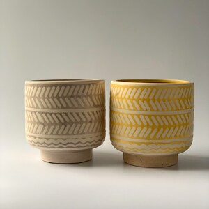 May include: Two ceramic planters with a geometric pattern. The planter on the left is a light gray color with a white pattern. The planter on the right is a yellow color with a white pattern.