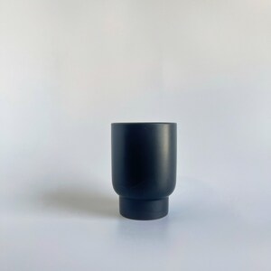 May include: A matte black ceramic planter with a cylindrical shape and a slightly flared base. The planter is designed for indoor or outdoor use and is suitable for various plants. The minimalist design complements modern decor.