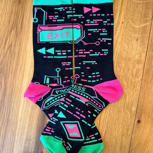 Könnte beinhalten: Ein Paar schwarze Socken mit einem leuchtend neonpinken und türkisfarbenen digitalen Schnittstellenmuster. Das Design enthält den Text EXIT und PROGRESS sowie verschiedene geometrische Formen, Linien und Symbole, die an einen Retro-Videospielbildschirm erinnern.