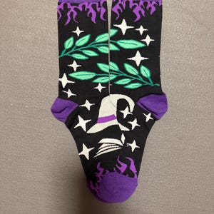 Könnte beinhalten: Ein Paar schwarze Socken mit einem violetten Flammenmuster am oberen Rand, grünen Blattzweigen, weißen vierzackigen Sternen und einem weißen Hexenhut mit violettem Band. Die Socken haben violette Bündchen, Fersen und Zehenbereiche.