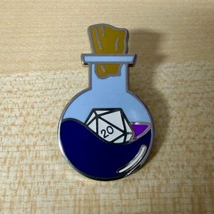 Könnte beinhalten: Runder Emaille-Pin mit einem Fläschchen-Design. Die Flasche ist hellblau mit einer dunkelblauen Flüssigkeit und einem weißen und schwarzen D20-Würfel im Inneren. Die Flasche hat einen Korken.