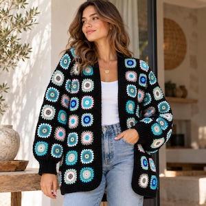 Könnte beinhalten: Eine langärmelige, vorne offene Strickjacke aus schwarzen Häkelquadraten, die jeweils ein kreisförmiges Muster in Blau-, Rosa-, Weiß- und Grautönen aufweisen. Das Kleidungsstück hat einen gerippten schwarzen Saum an der Vorderöffnung.