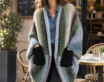 May include: An open-front, oversized knit cardigan featuring a variegated colour palette of green, blue, and purple tones. The garment includes two large, solid black patch pockets on the front and has a relaxed, cocoon-like silhouette with wide sleeves.