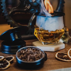 May include: A close-up of a glass of amber liquid with smoke rising from it. The glass is on a white coaster and there is a black lid on top of the glass. There are dried orange slices and a black metal container with dried herbs on a wooden surface.