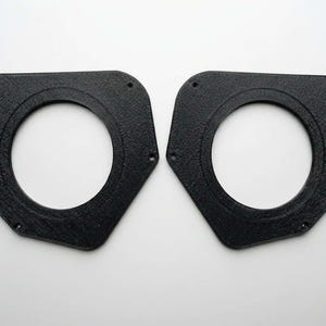 Könnte beinhalten: Zwei schwarze, texturierte, asymmetrische Lautsprecher-Adapterplatten mit einem zentralen kreisförmigen Ausschnitt und vier kleinen Befestigungslöchern, die um den Umfang jedes Teils angeordnet sind. Diese starren Komponenten haben eine matte Oberfläche.