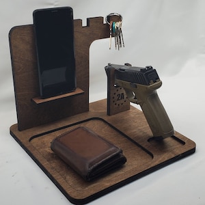 May include: A wooden organizer in a dark brown finish, designed to hold a smartphone, keys, wallet, and a handgun. The organizer features a slot for the phone, a key ring holder, a tray for the wallet, and a stand for the firearm. The text "2A" is visible.